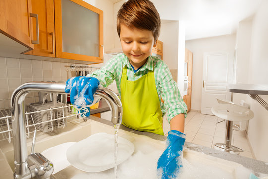 Happy kid boy rinsing dishes in the sink