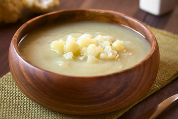 Cream of cauliflower soup in wooden bowl, photographed on wood with natural light (Selective Focus, Focus on the front of the cauliflower on the soup)