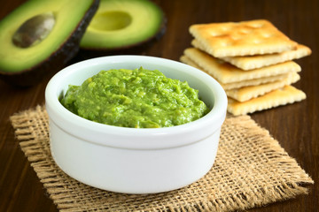 Fresh avocado cream or guacamole with soda crackers and half avocados in the back, photographed...