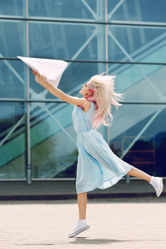 Outdoor full body portrait of young beautiful girl throwing big white paper airplane. Model with long blond, pink hair, wearing vintage dress, sneakers. Glass wall on background. Sunny day light.