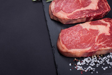 Two fresh raw marble meat, black Angus ribeye steak with spices on a dark stone background.