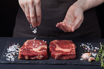 A female chef sprinkles sea salt with two fresh raw ribeye steaks from marbled beef on a dark background. Nearby is a mixture of peppers, garlic and rosemary