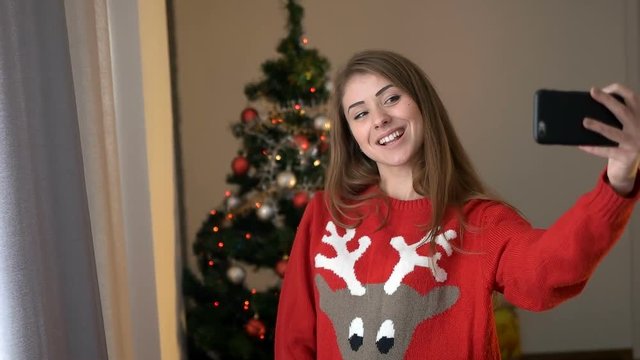 Young woman in sweater with geer taking selfie on smart phone on christmas tree background, she posing and smiling on camera.
