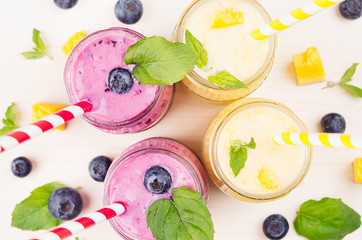 Freshly blended yellow and violet  fruit smoothie in glass jars with straw, mint leaves, mango slices and berry, close up, top view. Soft white wooden board background.