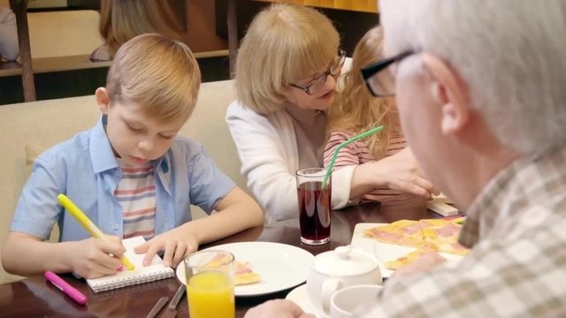 Grandparents and grandkids having lunch in the cafe: little boy drawing on paper and girl sitting on knees of grandma and building with toy bricks