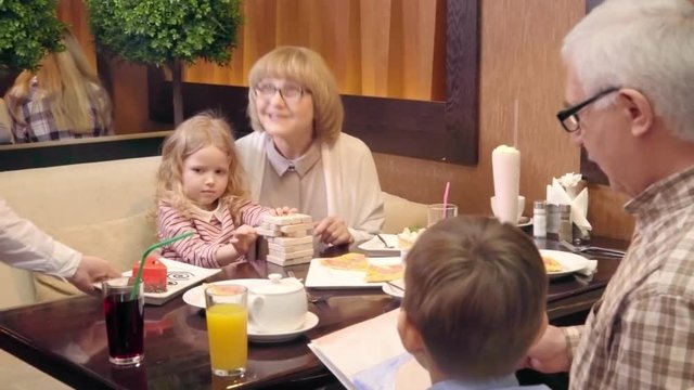 Tracking of grandparents with kids sitting at table in restaurant: grandmother building jenga tower with girl and granddad looking at drawing with grandson while waiter serving dessert