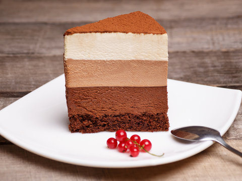 Slice Of The Three Chocolate Mousse Cake On A White Plate Against A Wooden Background