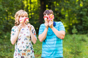 Fototapeta premium Pregnancy, family, happiness and fun concept - Man and pregnant woman have fun with candy in the park