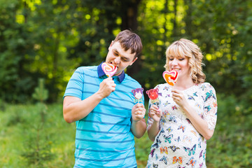 Fototapeta premium Pregnancy, family, happiness and fun concept - Man and pregnant woman have fun with candy in the park