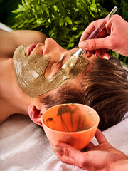 Mud facial mask of man in spa salon. Massage with clay full face. Girl on with therapy room. Man lying wooden spa bed. Beautician with bowl therapeutic procedure isolated background. Close-up of a man