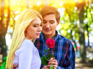 Fototapeta premium Spring couple walking and kissing summer park. Boyfriend and girlfriend walk in sun shine. First date of two young people . Guy gives girl red rose flower. Fairy-tale love at first sight.