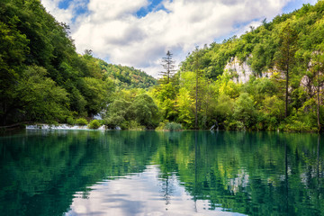 Fototapeta na wymiar Plitvice Lakes National Park beautiful daytime landscape, green forest, waterfalls, blue cloudy sky and reflection in amazing green water of nature lake, image suitable for wallpaper or guide book