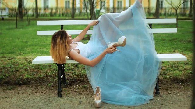 Attractive girl in white and blue dress lies on bench with bare feet and handles high heeld shoes posing during photo shoot.