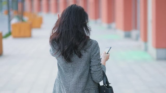 Back span of the young tender amazing business girlin strict gray jacket and black dress model walking on the urban busy street with phablet smartphone in her hand Steadicam slow motion shot