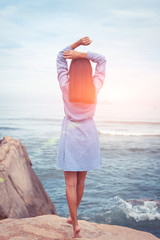 A sunny portrait of beautiful asian woman in dress back stand and rise hands on a stone by the sea