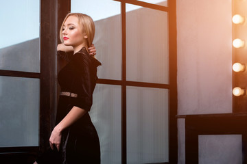 Studio shot of young and beautiful girl standing near window in black dress wearing in studio. Blonde girl