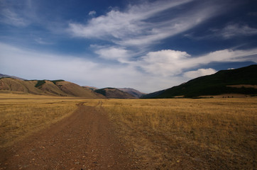 Fototapeta na wymiar Road through a dry desert steppe on a highland mountain plateau with yellow grass with ranges of hills on a horizon skyline under blue sky and white clouds, Kurai, Altai, Siberia, Russia