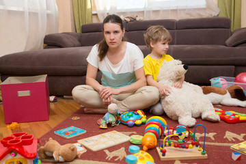 Young pregnant mother and her little son, at home. Mother and little son  sit on a floor. Mom tired to tidy up the house. Child scattered toys. Mess in the house. Mother has quarreled with the son.