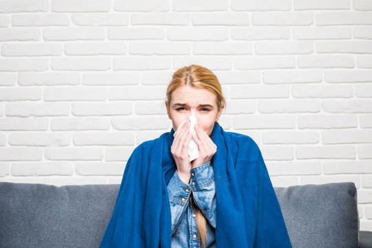blonde woman sneezing in a tissue on sofa in the living room