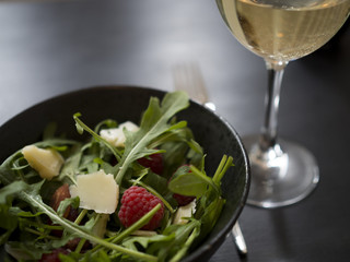 Fresh rocket salad with raspberries, roasted almonds and cheese in black ceramic bowl, and a glass of white wine on black wooden table