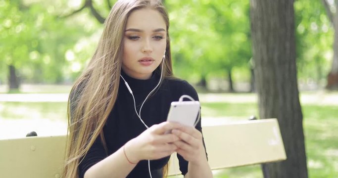 Pretty young woman browsing smartphone and listening to music with headphones while sitting on bench in park