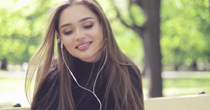 Portrait of beautiful young woman with headphones smiling at camera while sitting on bench in park with green trees