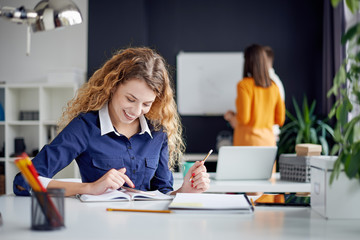 Young happy people discussing business standing at the whiteboard in modern light office, beautiful woman sitting at the table at forefront