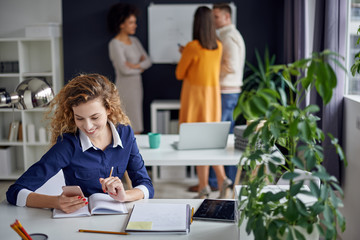 Young happy people discussing business standing at the whiteboard in modern light office, beautiful woman sitting at the table at forefront