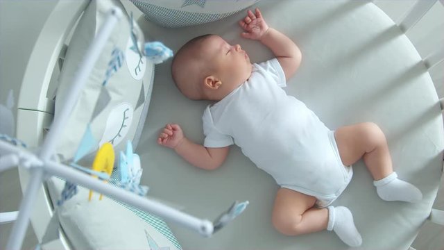 newborn lies in the round white bed with mobile and toys