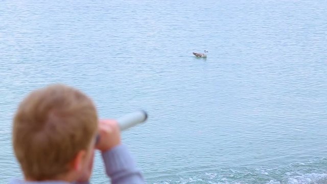 Little child looks through old telescope at something interesting in blue sea. Happy 8 year old kid has fun on beach on vacation in cool spring or autumn season. Real time hd video footage.