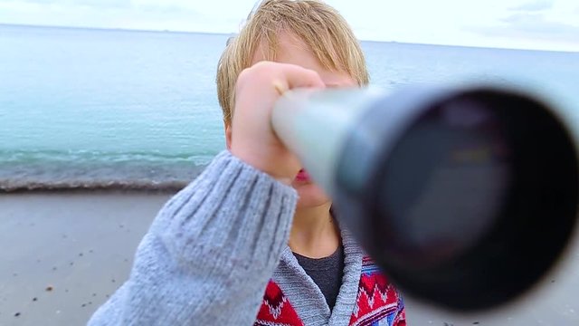 Little child looks through old telescope at something interesting in blue sea. Happy 8 year old kid has fun on beach on vacation in cool spring or autumn season. Real time hd video footage.