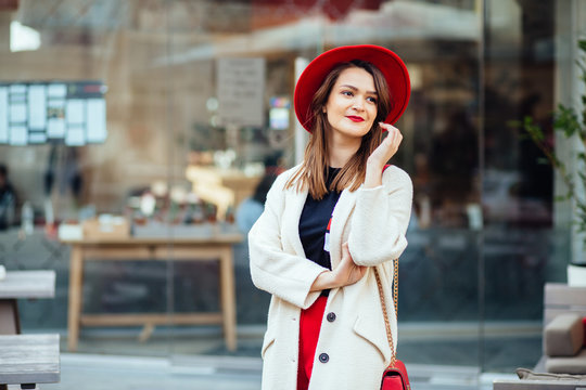 Portrait of positively a stylish brunette business woman in red hat with a red handbag wearning in white coat , red trousers waiting in front of hotel or boutique window.