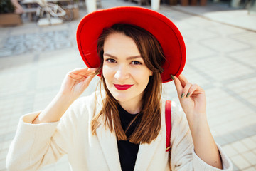 Pprtrait of young positively woman traveler in bright red hat and white coat looking at camera in...