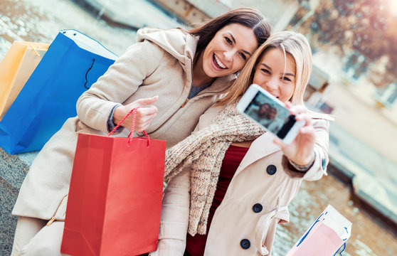 Shopping time. Beautiful smiling girls enjoying in shopping and taking a selfie with mobile phone. Consumerism, fashion, lifestyle concept