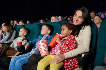 Cute little girl sipping her drink sitting on the lap of her mother at the cinema enjoying watching...