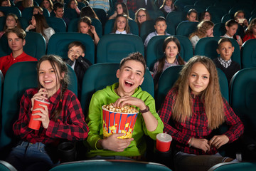 Young boy laughing cheerfully enjoying a funny movie at the cinema with his friends eating popcorn...