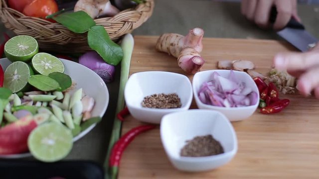 Female hand crush garlics and slice them with chef knife on bamboo wooden chopping block with other Thai herbs beside for making Thai red curry or Panang recipe.