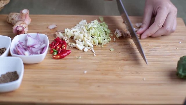 Female hand slice galanga with chef knife on bamboo wooden chopping block with other Thai herbs beside for making Thai red curry or Panang recipe.