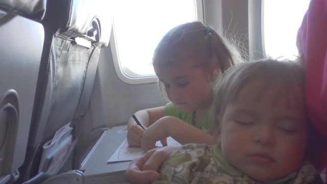 Little girl child draws,  sitting on inside airplane. Baby boy is sleeping with mom in her arms.