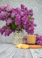 glass coffee Cup with milk on a wooden stand with cookies on a background of purple lilac bouquet