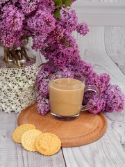 Fototapeta na wymiar glass coffee Cup with milk on a wooden stand with cookies on a background of purple lilac bouquet