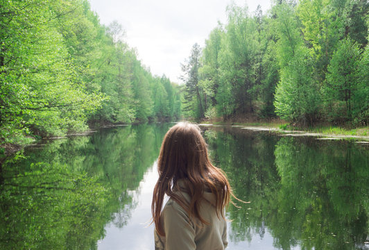 Girl Stands With His Back To The Camera. Against The Background Of A Beautiful River. Green Forest