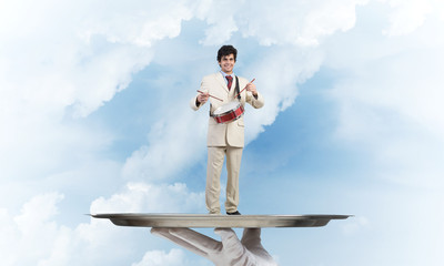 Young businessman on metal tray playing drums against blue sky background