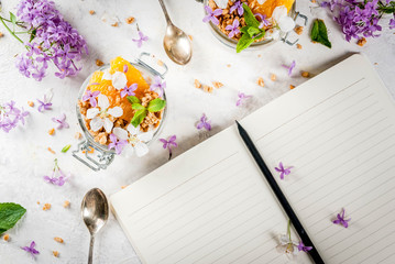 Trendy exotic food. Vegan paleo diet. Breakfast. Yoghurt with granola, slices of orange, mint and edible flowers  lilac, cherry. On white stone table, with notebook and ingredient, Copy space top view
