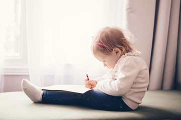 Portrait of cute adorable white Caucasian little girl drawing with pencil on paper, sitting on couch indoor, looking serious concentrated,  lifestyle childhood early development