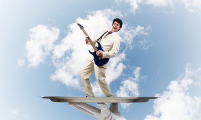 Businessman on metal tray playing electric guitar against blue sky background