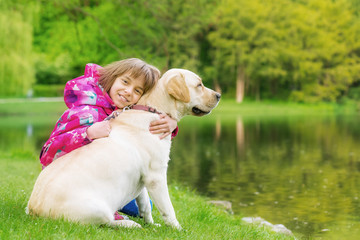 Little girl with labrador retriever on walk in park. Child sitting on green grass with dog -...