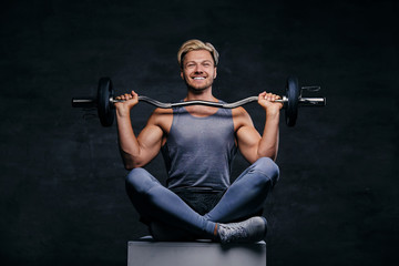 Obraz na płótnie Canvas Smiling blond sporty male sits on a white box with crossed legs and holds barbell.