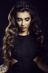Fototapeta premium Portrait of gorgeous young model with dark long silky wavy hair, thick eyelashes and full lips wearing lace dress and jewel crown standing on black background with her eyes down. Studio shot