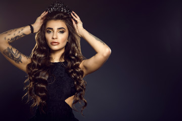 Portrait of gorgeous glam tattooed model with long wavy silky shiny hair and provocative make up putting up a jewel crown on her head. Isolated on black background with backlight. Copy-space © obrik